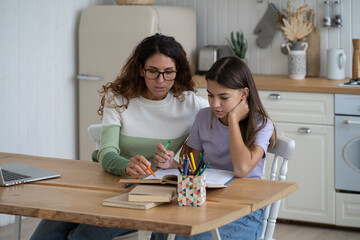 Diligent pensive teenage girl sits next to mother helping to complete tasks of school teacher. Caring attentive millennial woman sits with daughter at kitchen table with textbooks and notebooks