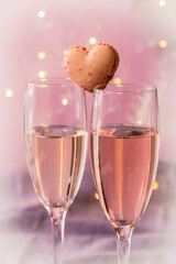 Two glasses with sparkling wine with a heart shaped macaron in between, pink pastel background with fairy lights, romantic atmosphere, vertical