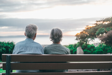 Head shot close up portrait happy grey haired middle aged woman with older husband, enjoying sitting on bench at park. Bonding loving old family couple embracing, looking sunset..