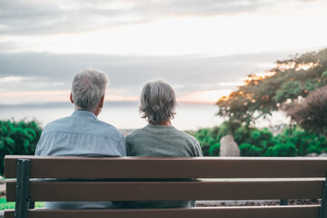 Head shot close up portrait happy grey haired middle aged woman with older husband, enjoying sitting on bench at park. Bonding loving old family couple embracing, looking sunset..