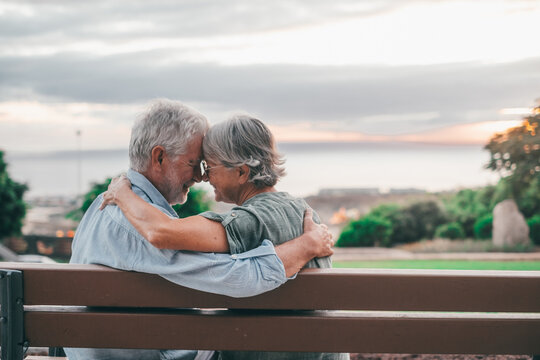 Head shot close up portrait happy grey haired middle aged woman snuggling to smiling older husband, enjoying sitting on bench at park. Bonding loving old family couple embracing, looking sunset..
