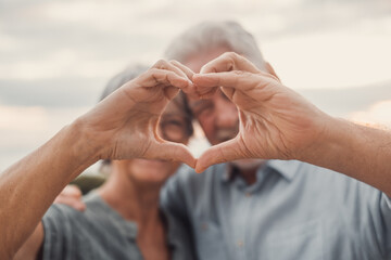 Close up portrait happy sincere middle aged elderly retired family couple making heart gesture with fingers, showing love or demonstrating sincere feelings together outdoors, looking at camera..