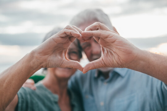 Close up portrait happy sincere middle aged elderly retired family couple making heart gesture with fingers, showing love or demonstrating sincere feelings together outdoors, looking at camera..