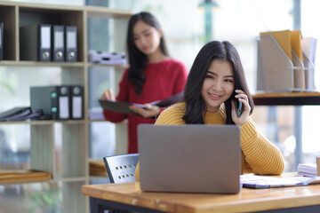 Attractive young Asian businesswoman working with laptop at her desk and having coworkers in the office.