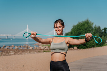 Fit brunette hispanic girl in sportswear stretching rubber tapes, doing workout at beach with view on bay, bridge on summer sunny day. Cheerful hispanic young woman training outdoors. Fitness.