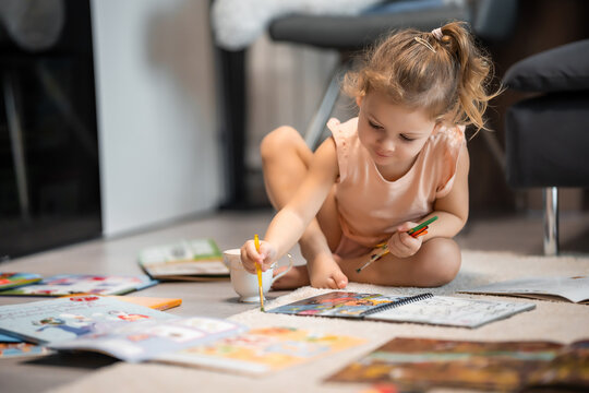 Little girl sits on the floor at home and draws with paints and brushes in a coloring book. Early childhood creativity and education
