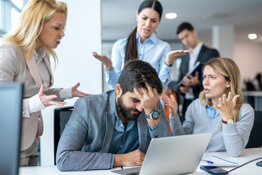 Frustrated businessman covering face with hand, having serious problems while his co-workers making him feels guilty for not doing his job properly at office
