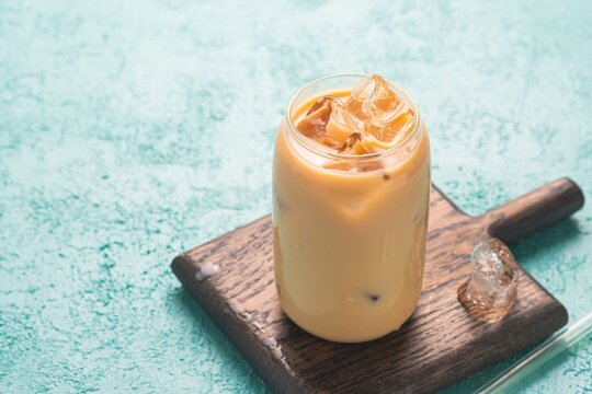 Refreshing drink, Hong Kong style milk tea with ice cubes in a jar-shaped glass cup on a brown board on a turquoise concrete background. Refreshing drinks