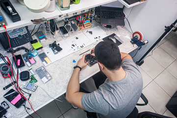 Electronics repair shop, a repairman is surrounded by tools and equipment. A technician repairs, cleans, controls a smartphone. Workplace top view, close-up.