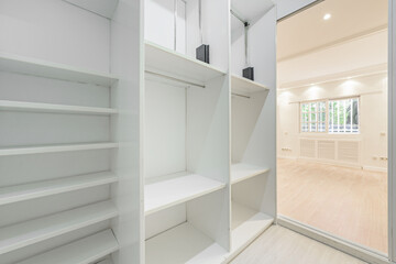 An empty room with white painted walls with access to a dressing room with multiple white marera shelves and French style aluminum windows