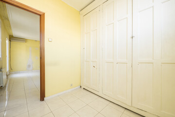 An empty room with walls covered by a large built-in wardrobe with vintage-style white wooden doors and yellow painted walls, a white stoneware floor and a door to another room