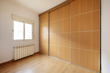 An empty room with walls covered by a large built-in wardrobe with four sliding doors made of oak wood and aluminum edges and a window with an aluminum radiator