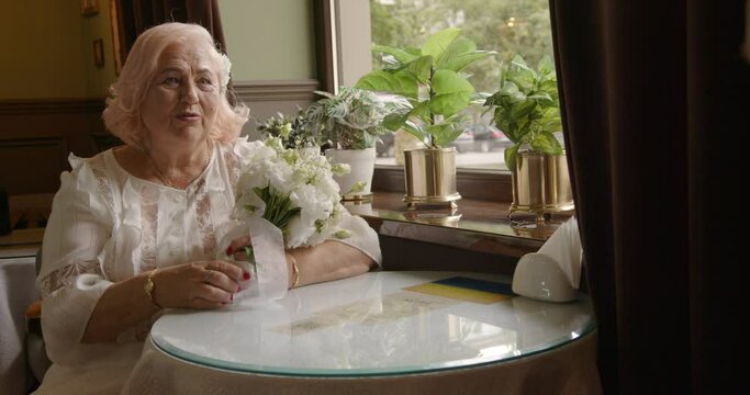 A 70-year-old bride sits at a table with an elderly groom with a bouquet of white flowers and talks. old couple on a date. fat old beautiful bride.