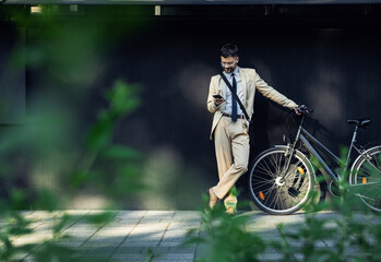 Portrait of business man with bicycle standing in front of office building looking at smartphone.