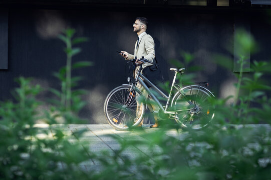 Portrait of business man with bicycle walking in front of office building using smartphone.