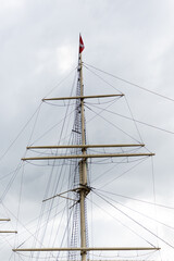 Takelage eines großen Segelbootes mit roter Flagge vor dunklen Wolken in Hamburg.
