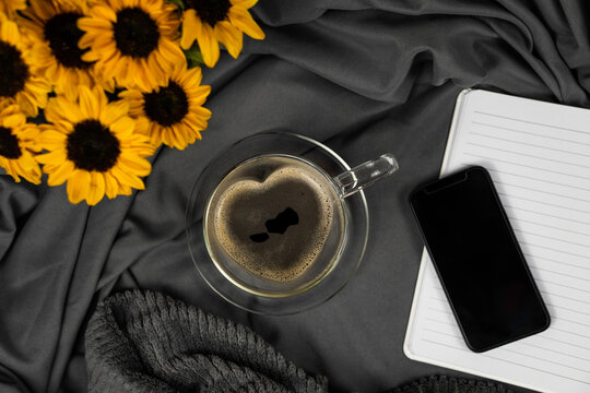Herzförmige Glastasse mit Kaffee gefüllt auf Glasunterteller auf grauem Satinstoff, kleine Sonnenblumen teilweise links im Bild, rechts noch teilweise schwarzes Smartphone, Notizheft, Topview, horizon