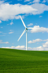 A single wind turbines slowly turning rises above green hilly farming fields with blue sky and puffy clouds.