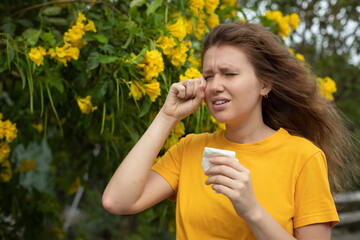 Portrait of beautiful young allergic woman is suffering from pollen allergy or cold on natural flower flowering tree background at spring or sunny summer day sneezes, blowing her runny nose rubs eyes