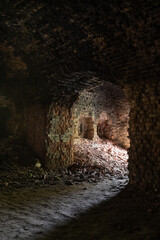 A long dark dimly tunnel of an abandoned military bunker or bomb shelter with broken red brick walls. The arched ceiling is illuminated by daylight from the outside. The floor is dirty and dusty