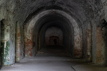 Fototapeta na wymiar A long dark dimly tunnel of an abandoned military bunker or bomb shelter with broken red brick walls. The arched ceiling is illuminated by daylight from the outside. The floor is dirty and dusty