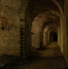 Naklejka premium A long dark dimly tunnel of an abandoned military bunker or bomb shelter with broken red brick walls. The arched ceiling is illuminated by daylight from the outside. The floor is dirty and dusty