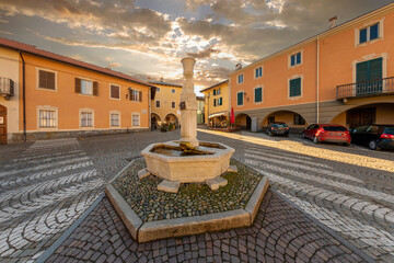 Obraz premium Peveragno, Cuneo, Italy - January 09; 2023: marble fountain with drinking trough on cobblestones in Piazza Santa Maria with ancient palaces decorati with arcade under cloudy sky at sunset