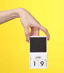 Woman's hand holds wooden calendar with date june 19 on a yellow background