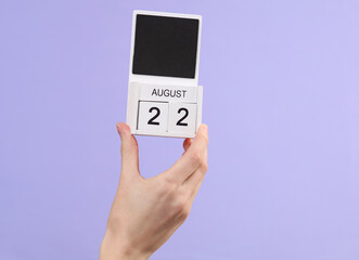 Woman's hand holds wooden calendar with date august 22 on pastel purple background