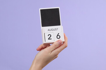 Woman's hand holds wooden calendar with date august 26 on pastel purple background