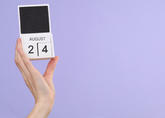 Woman's hand holds wooden calendar with date august 24 on pastel purple background