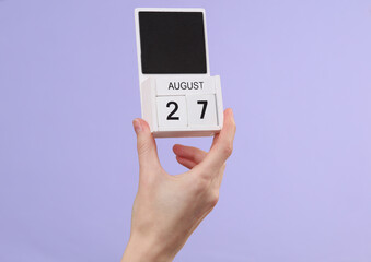 Woman's hand holds wooden calendar with date august 27 on pastel purple background