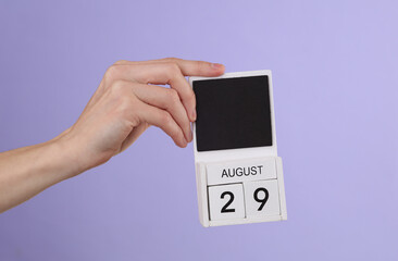 Woman's hand holds wooden calendar with date august 29 on pastel purple background