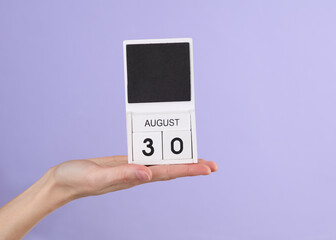 Woman's hand holds wooden calendar with date august 30 on pastel purple background