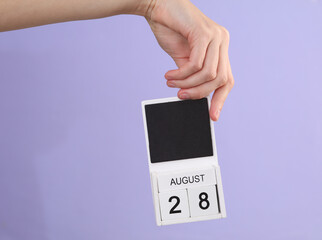 Woman's hand holds wooden calendar with date august 28 on pastel purple background