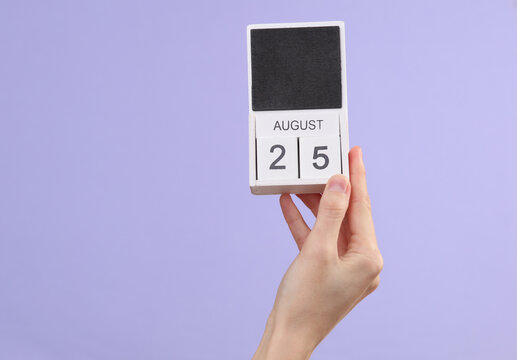 Woman's hand holds wooden calendar with date august 25 on pastel purple background