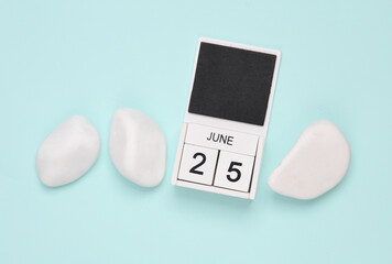 White wooden calendar with the date june 25 on blue background with white pebbles