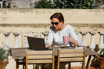 Woman using a laptop and drinking coffee while sitting outdoors in terrace.