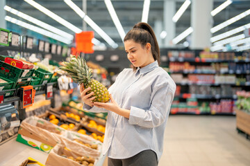 Woman buying fresh fruits in the supermarket, choosing healthy treats, shopping pineapples. Young woman standing in a supermarket, looking at fresh juicy pineapple.