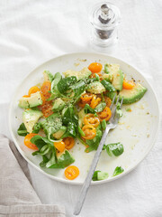 Colorful mixed salad of orange cherry tomatoes, avocado and lamb's lettuce. Plate with fork on table cloth next to pepper grinder.