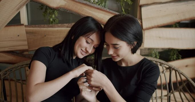 Couple, lgbt and lesbian women at home.Low angle handheld shot of young lesbian couple smiling and touching hands gently while sitting on sofa against window in light living room at home. LGBT concept