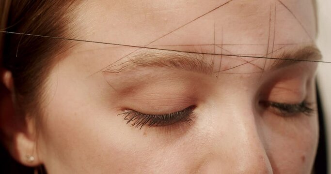 Crop cosmetician making creative makeup. Closeup handheld shot of crop makeup artist in latex gloves using thread to stamp lines on forehead of woman in salon.