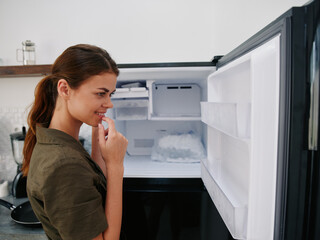 Fototapeta na wymiar Woman smiling with teeth looking into camera in kitchen at home opened freezer empty with ice inside, home refrigerator, defrosted, view from back, stylish interior.