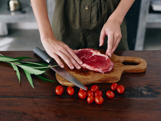 Woman with knife in hand cutting fresh steak meat for roasting in kitchen with salt pepper and other spices on table, red cherry tomatoes and herbs, preparing dinner. wooden table, top view.