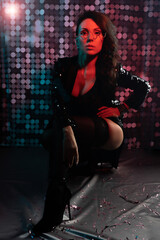 Fototapeta na wymiar A gorgeous brunette in a black bodysuit, black stockings and a sequined jacket at a party. Night club. Red backlight in the dark. Photo with colored light.