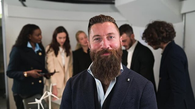 close up hipster businessman posing looking at camera smiling and nod with head.  Defocused teamwork working behind him. Successful leadership concept.