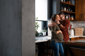 Handsome man with an adorable pregnant woman embracing and smiling while bonding together at home. Happy family looking forward to the birth of their new baby. Spending time together in the kitchen.