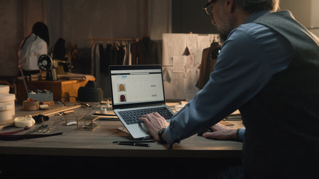 Successful man browsing personal clothing shop on laptop or watching orders statistic. Tailor sits by the table at luxury designer atelier and works with clothing range on website. Concept of fashion.