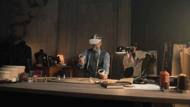 Mature tailor wearing VR headset with wireless controllers, sits at the table in luxury designer atelier, gesturing and creates design in virtual reality. Concept of technologies of future in business