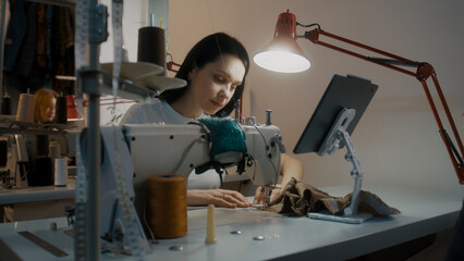 Female fashion designer or dressmaker touches tablet, sits at sewing machine and tailoring suit. Seamstresses working on custom apparel at background. Atelier or sewing workshop. Fashion concept.